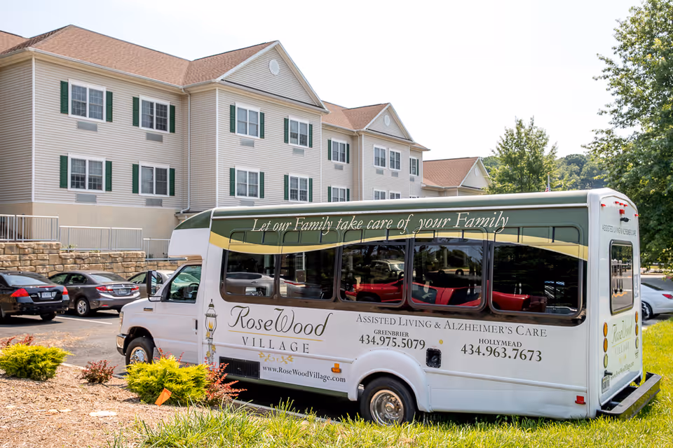 A white shuttle bus parked outside a multi-story assisted living facility building with beige siding and green window shutters. The bus has signage for RoseWood Village Assisted Living & Alzheimer's Care with contact numbers and the slogan 'Let our Family take care of your Family.' There are several cars parked nearby and trees in the background.