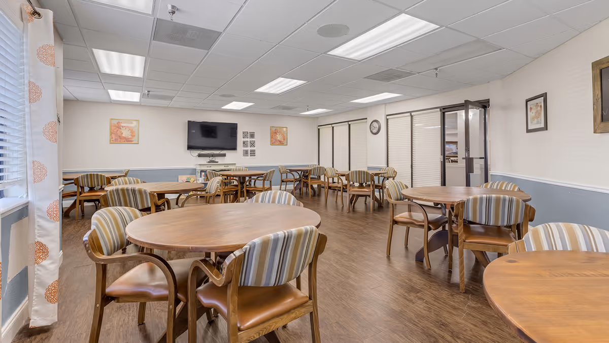 A spacious dining room with multiple round wooden tables and striped cushioned chairs arranged neatly. The room has wood flooring, a wall-mounted TV, framed artwork on the walls, and large windows with blinds and patterned curtains allowing natural light to enter.
