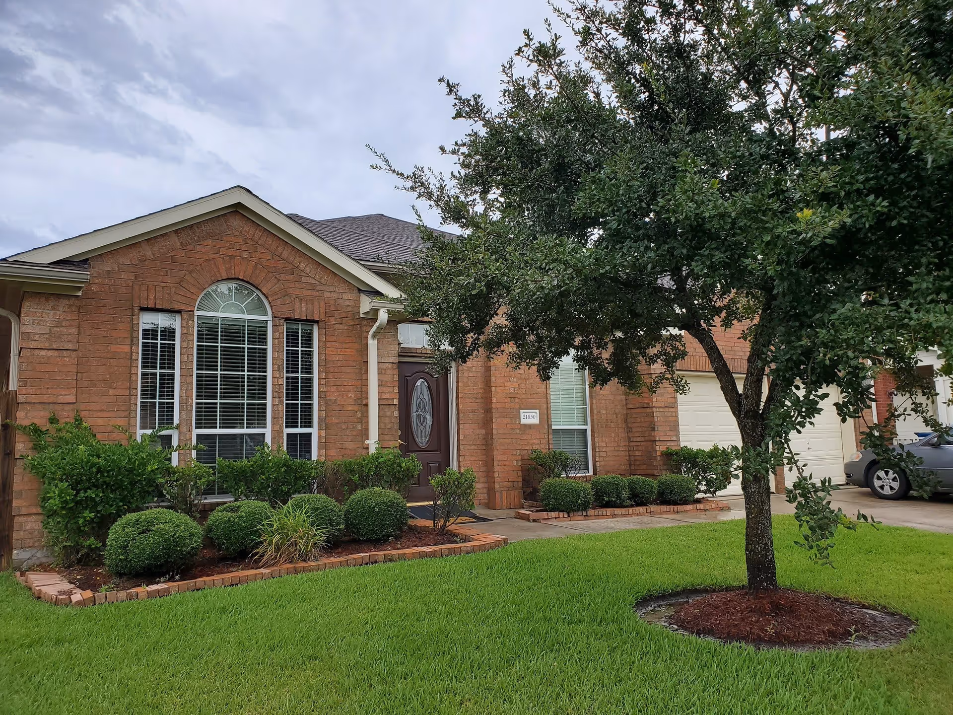Front exterior view of a brick residential building with a large window, a brown front door with decorative glass, neatly trimmed bushes, a green lawn, and a tree in the foreground. A driveway with a parked car is visible on the right side.
