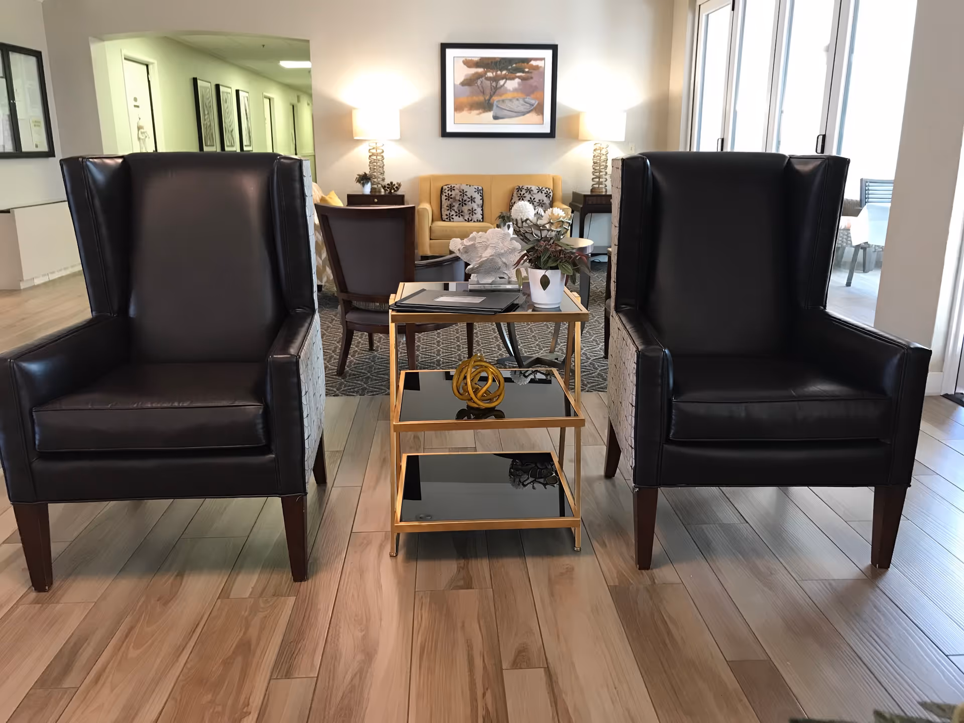 A cozy seating area in a senior living facility with two black leather armchairs facing forward and a small three-tiered glass and gold metal table between them. The table holds decorative items including a small plant and a sculpture. In the background, there is a yellow sofa with patterned cushions, two side tables with lamps, a framed painting on the wall, and a hallway with framed pictures on the walls. Large windows on the right let in natural light.