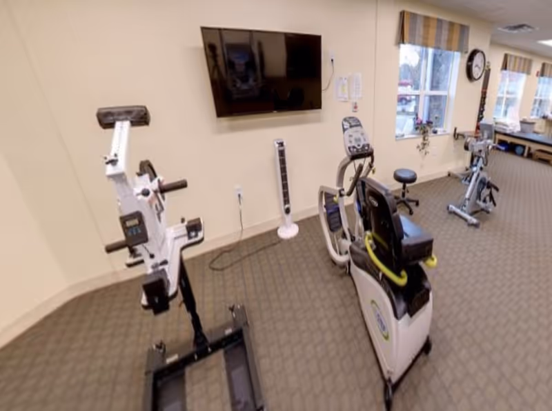 A carpeted exercise room with several stationary and recumbent bikes, a wall-mounted TV, and windows along one wall.