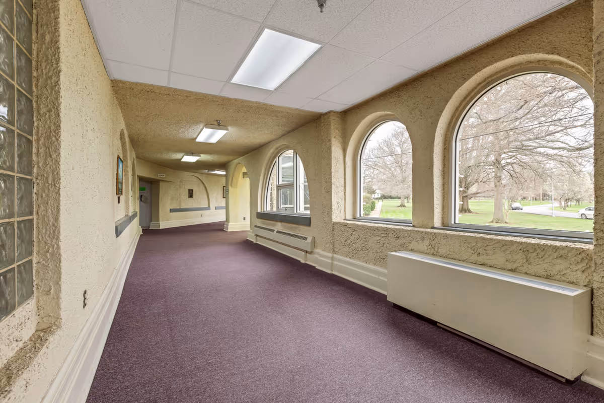 A long indoor hallway with textured beige walls and purple carpet. The hallway features large arched windows on the right side, allowing natural light to enter and providing a view of trees and a road outside. Fluorescent ceiling lights illuminate the space, and there are heating units below the windows.