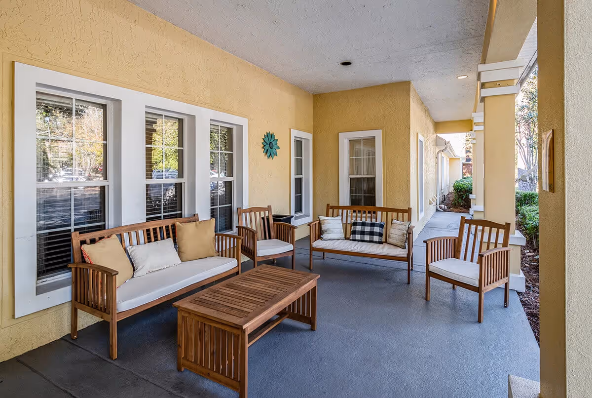 Covered exterior porch with wooden benches, chairs, cushions, and a coffee table along a yellow stucco building.