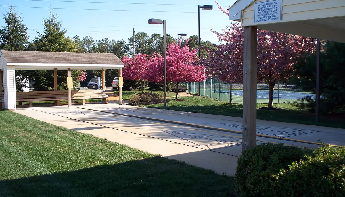 Outdoor recreational area with a covered shuffleboard court, benches, and blooming pink trees along a fenced tennis court in the background under a clear sky.