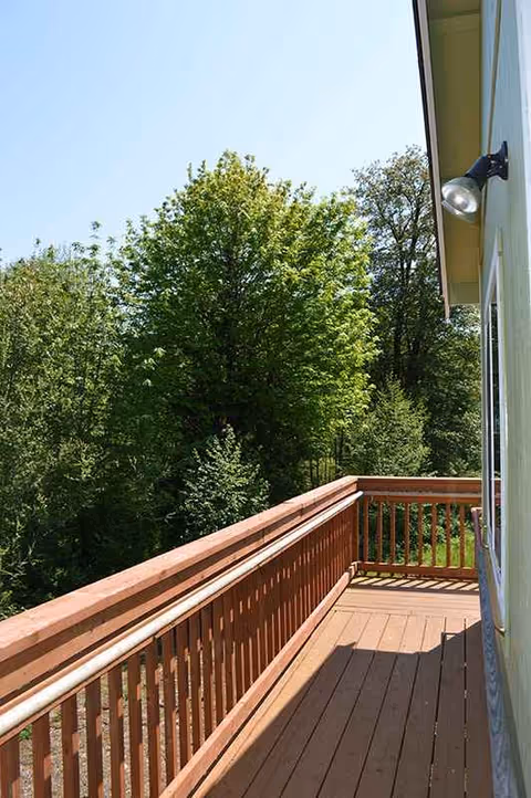 Wooden balcony extending from the building with a railing, overlooking green trees in bright outdoor light.