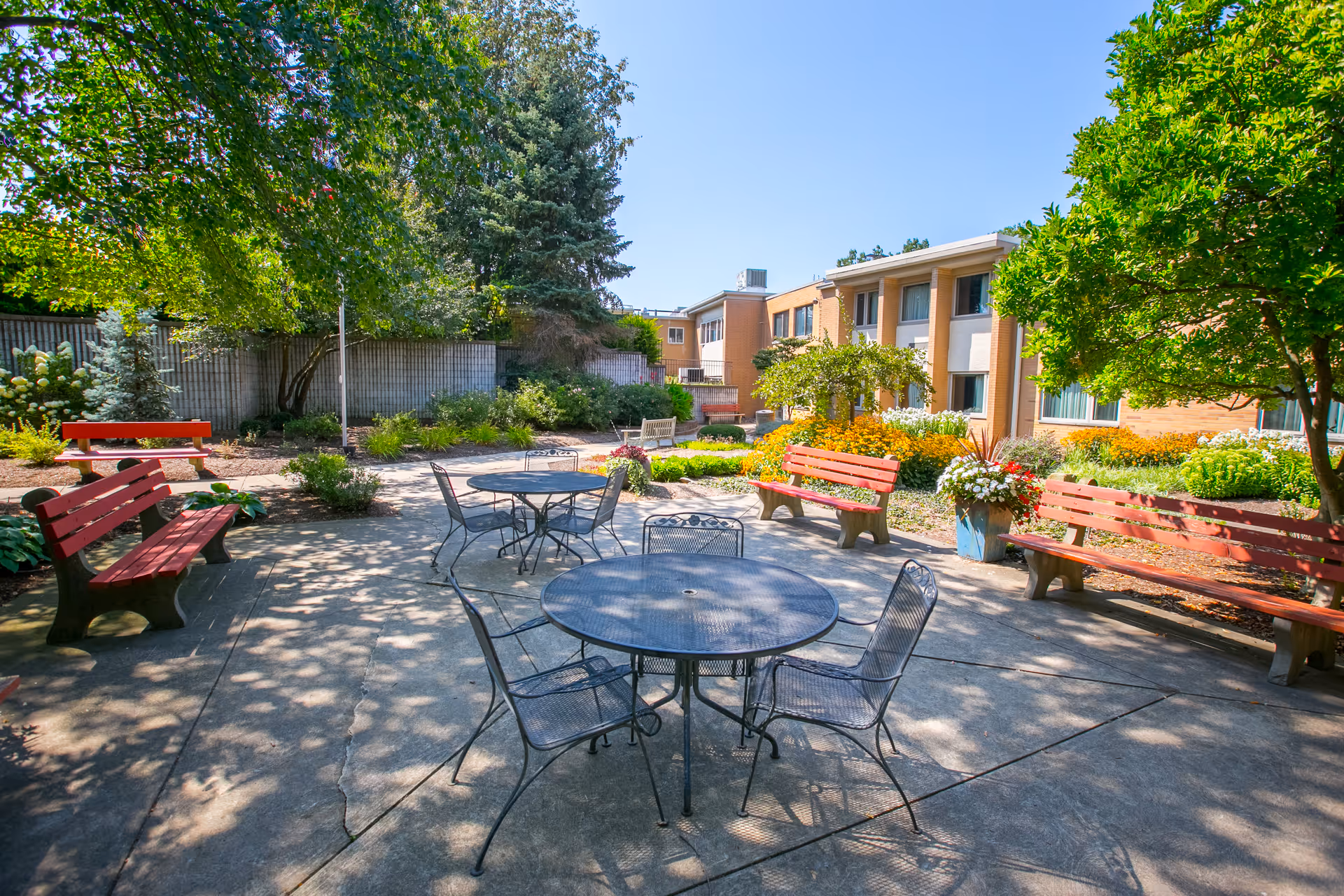 Outdoor courtyard area at Broadview Multi-Care Center featuring several red wooden benches, round metal tables with chairs, surrounded by trees, shrubs, and colorful flowers under a clear blue sky.