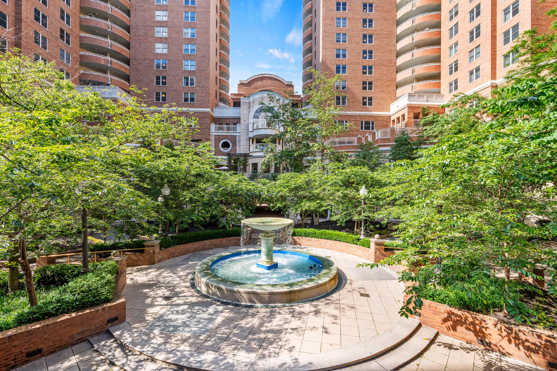 A courtyard with a circular water fountain in the center, surrounded by lush green trees and bushes. The courtyard is enclosed by tall brick residential buildings with balconies and numerous windows under a blue sky with some clouds.