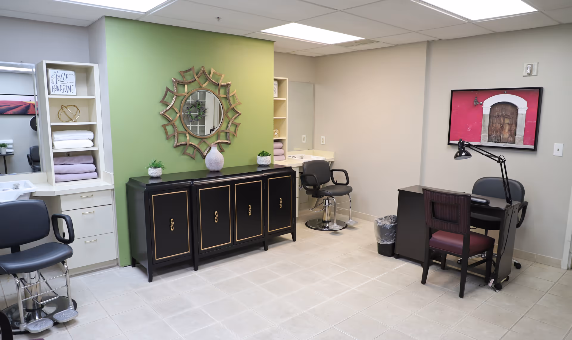 A well-lit room with a green accent wall featuring a decorative round mirror above a black cabinet. The room contains two black salon chairs, a small desk with a chair and a desk lamp, a sink, and shelves with neatly folded towels and decorative items. A framed picture with a red background and a wooden door hangs on the beige wall.