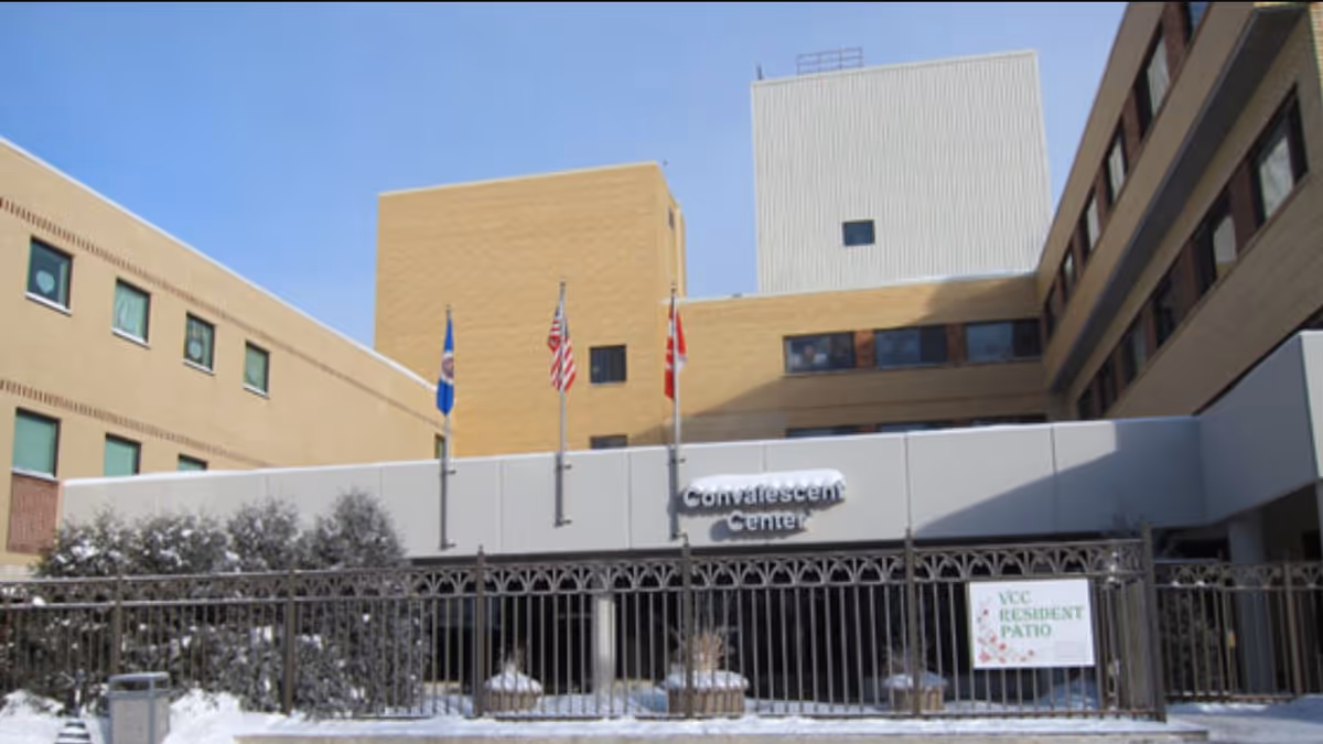 Front entrance of a convalescent care center building with flags and a fenced resident patio in light snow.