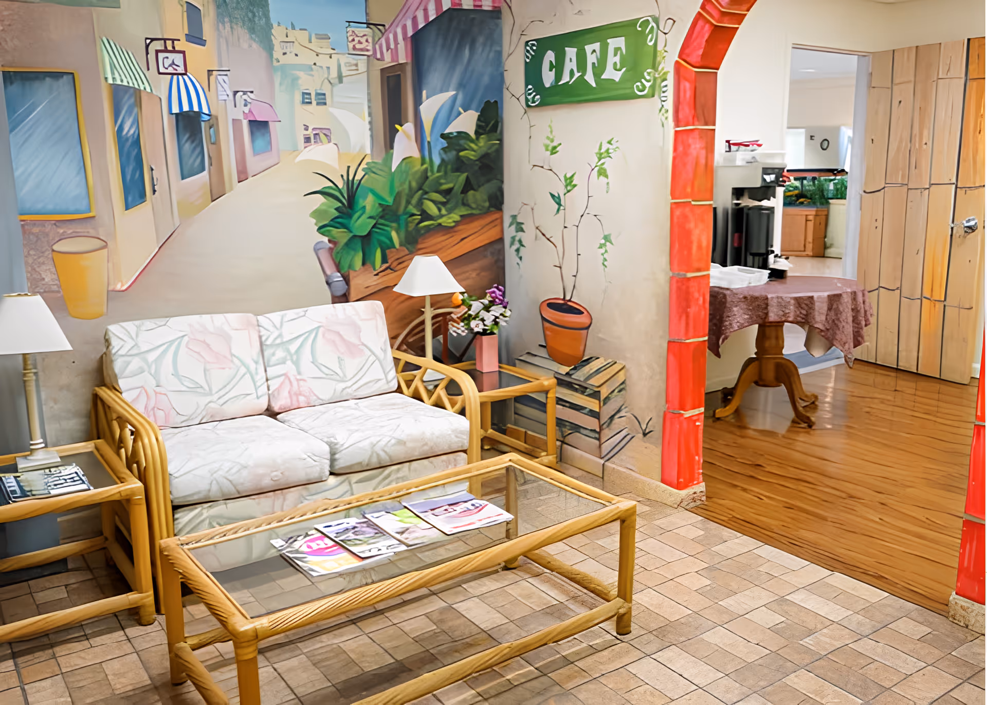 A cozy seating area with a floral-patterned couch, glass-top coffee table with magazines, and side tables with lamps. The walls are decorated with a mural depicting a street scene with plants and a green sign that says 'CAFE'. An arched doorway with red trim leads to another room with wooden flooring and a table covered with a brown tablecloth.