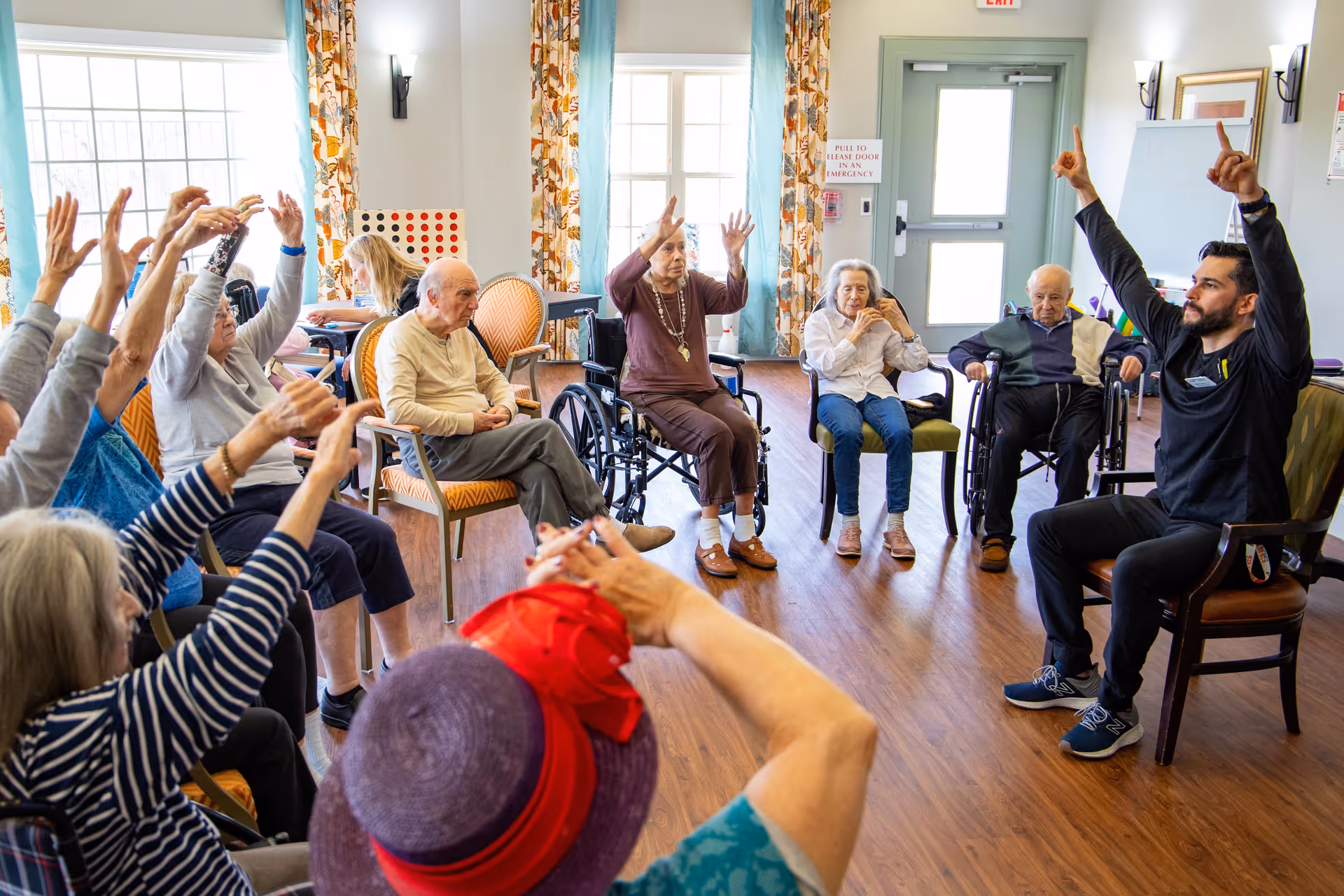 A group of elderly people seated in a circle in a bright room participating in a seated exercise session led by a male instructor. Some participants are in wheelchairs, and others are sitting on chairs, all raising their arms. The room has large windows with patterned curtains and wooden flooring.