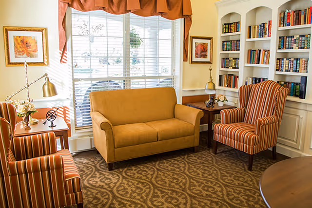 A cozy living room with a mustard yellow loveseat, two striped armchairs, a patterned carpet, built-in white bookshelves filled with books, two side tables with lamps and flowers, and a large window with blinds and a brown valance.