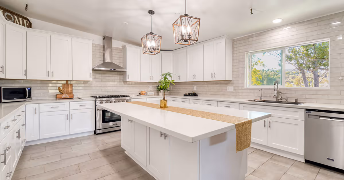 Bright and spacious kitchen with white cabinetry, a large central island with a beige runner, stainless steel appliances including a stove, range hood, microwave, and dishwasher. Two modern pendant lights hang above the island, and a window above the sink shows trees outside.
