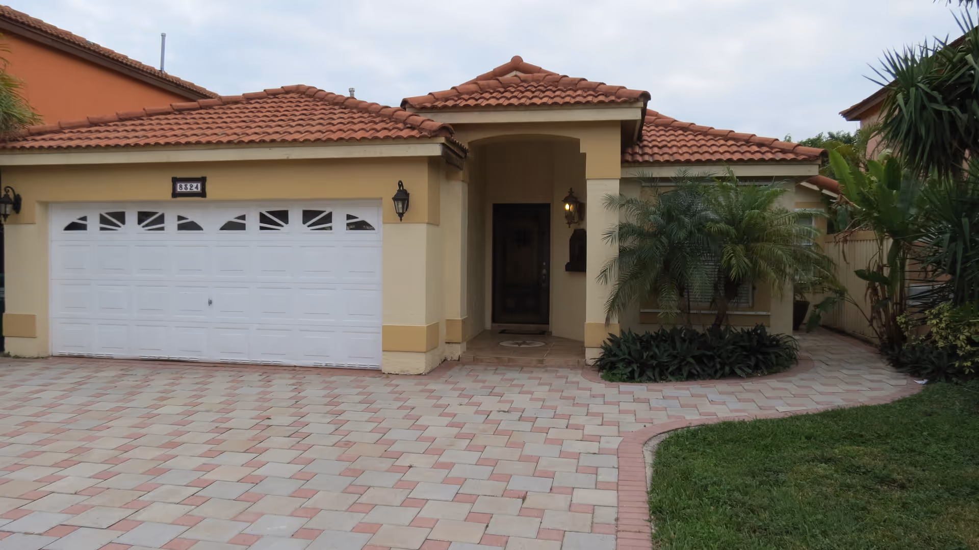 Front exterior view of a single-story house with a tiled roof, a double white garage door, a small covered entrance with a door, and surrounding greenery including palm plants and a lawn.