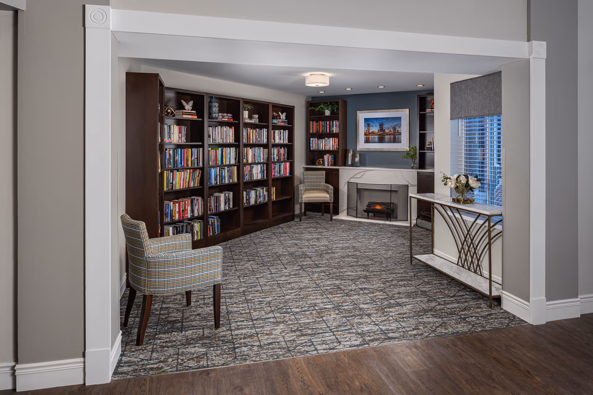 A cozy senior living facility library room with dark wooden bookshelves filled with books, two upholstered armchairs, a fireplace with a framed cityscape photo above it, a window with blinds and a decorative console table with a vase of flowers. The room has patterned carpet and wood flooring at the entrance.