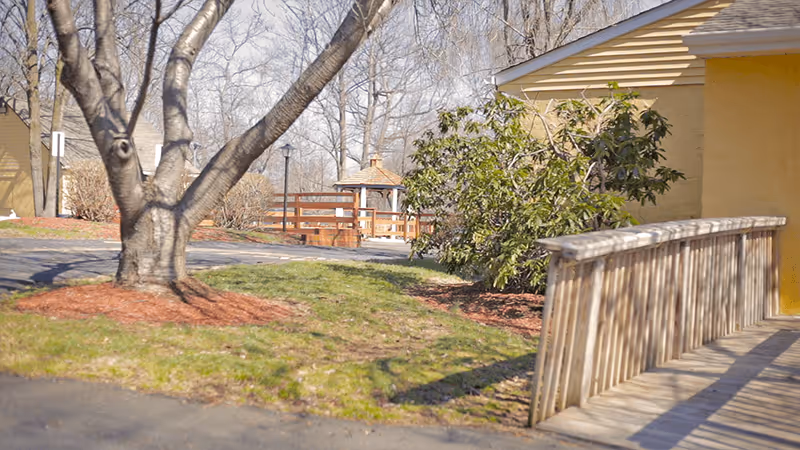 Outdoor view of a senior living facility area with a wooden ramp leading to a yellow building, a tree with bare branches, green grass, bushes, and a wooden gazebo in the background under a clear sky.