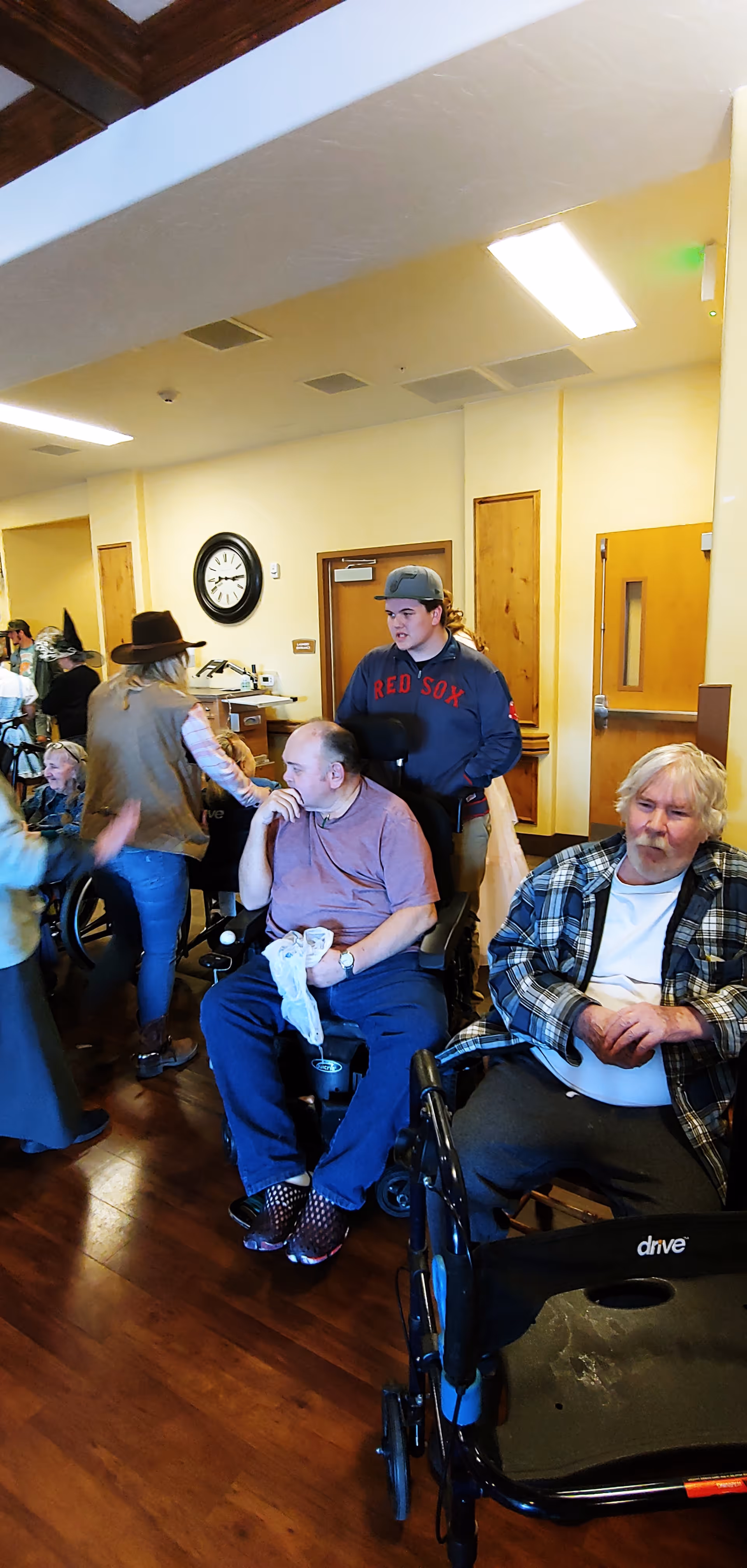 A group of elderly people and caregivers in a common area of a senior living facility. Some individuals are seated in wheelchairs, while others are standing and interacting. The room has wooden floors, beige walls, a clock on the wall, and wooden doors.