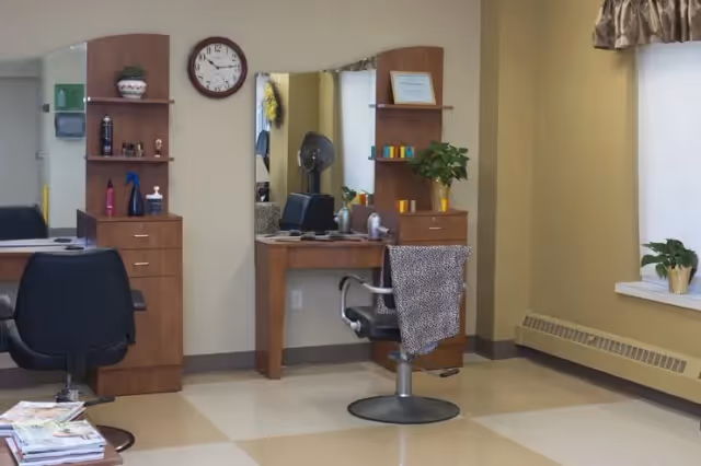 Interior view of a salon area in an assisted living facility with two black salon chairs, wooden shelving units with hair care products, a large mirror, a clock on the wall, and a window with a valance. One chair has a leopard print cloth draped over it.