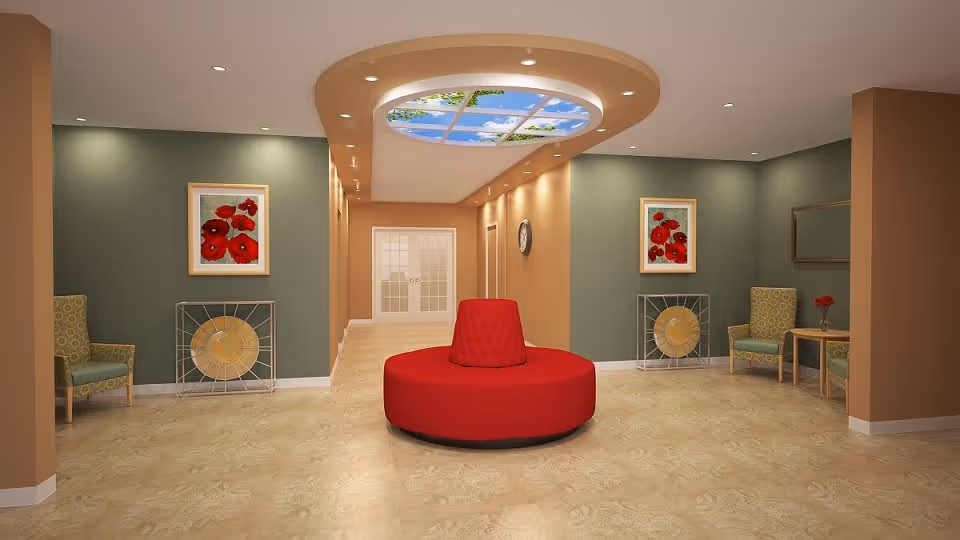 A spacious and well-lit interior common area with beige tiled flooring and walls painted in muted green and tan. The center features a round red cushioned bench with a backrest. On either side of the room, there are green patterned armchairs, small wooden tables with red flowers, and framed artwork of red flowers on the walls. The ceiling has a unique recessed oval design with a sky and tree canopy image, illuminated by recessed lighting. At the far end, double glass doors lead to another area.