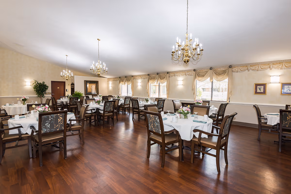 A spacious dining room with multiple round tables covered with white tablecloths, each set with plates, cups, and floral centerpieces. The room features wooden flooring, large windows with beige curtains, and elegant chandeliers hanging from the ceiling. The walls are light-colored with framed artwork and wall sconces providing additional lighting.