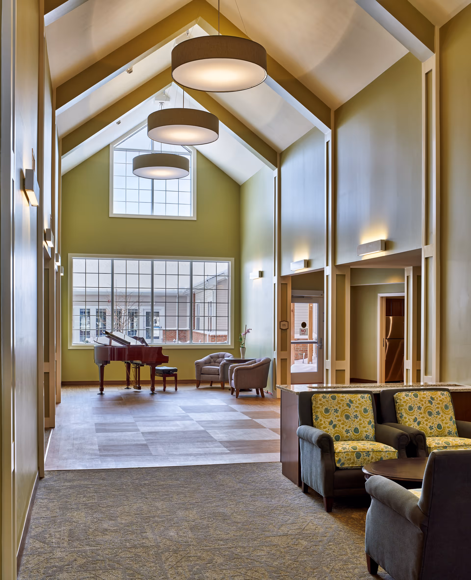 A spacious, well-lit common area with high vaulted ceilings and large windows. The room features a grand piano near the windows, two armchairs with a small table between them, and a seating area with two patterned armchairs and a small round table in the foreground. Modern circular pendant lights hang from the ceiling, and the walls are painted a soft green color.