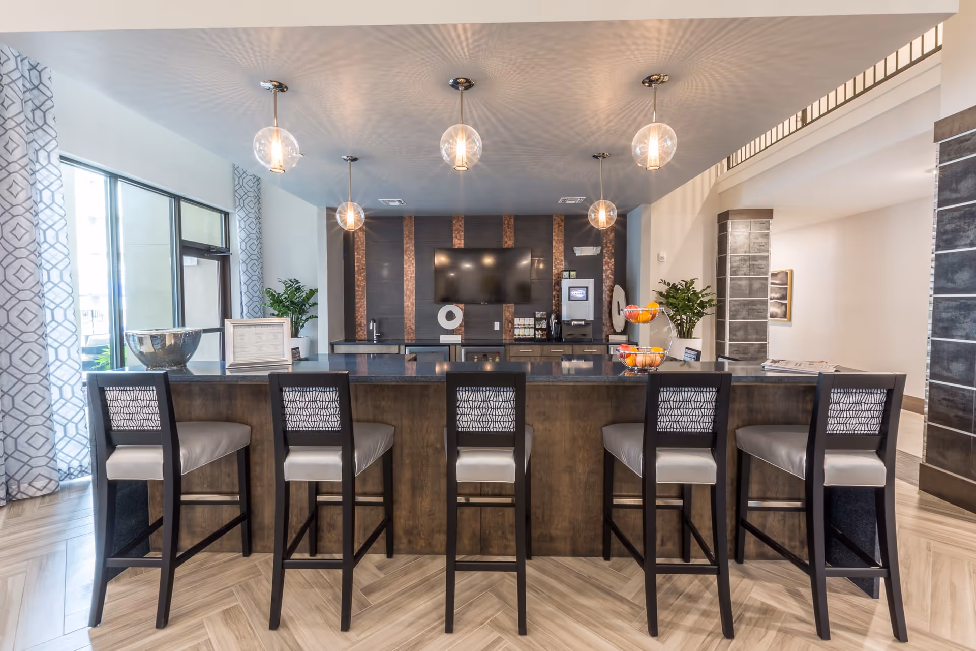 A modern kitchen area with a long island counter featuring five high chairs with patterned backs and cushioned seats. Above the island, five pendant lights with clear glass globes hang from the ceiling, casting decorative shadows. Behind the island is a wall with a mounted flat-screen TV, a coffee machine, a sink, and decorative elements. Large windows with patterned curtains are on the left side, and there are plants and a fruit basket on the counter.