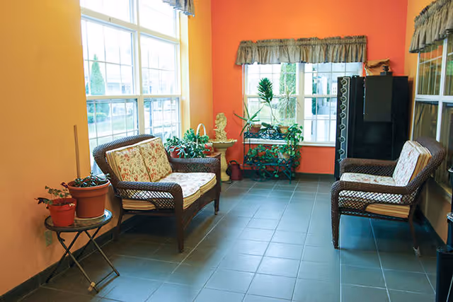 A cozy indoor seating area with two wicker chairs and a wicker loveseat, all with floral cushions. The room has orange and yellow walls, large windows with valances, several potted plants, and a small table with more plants. The floor is tiled in gray.