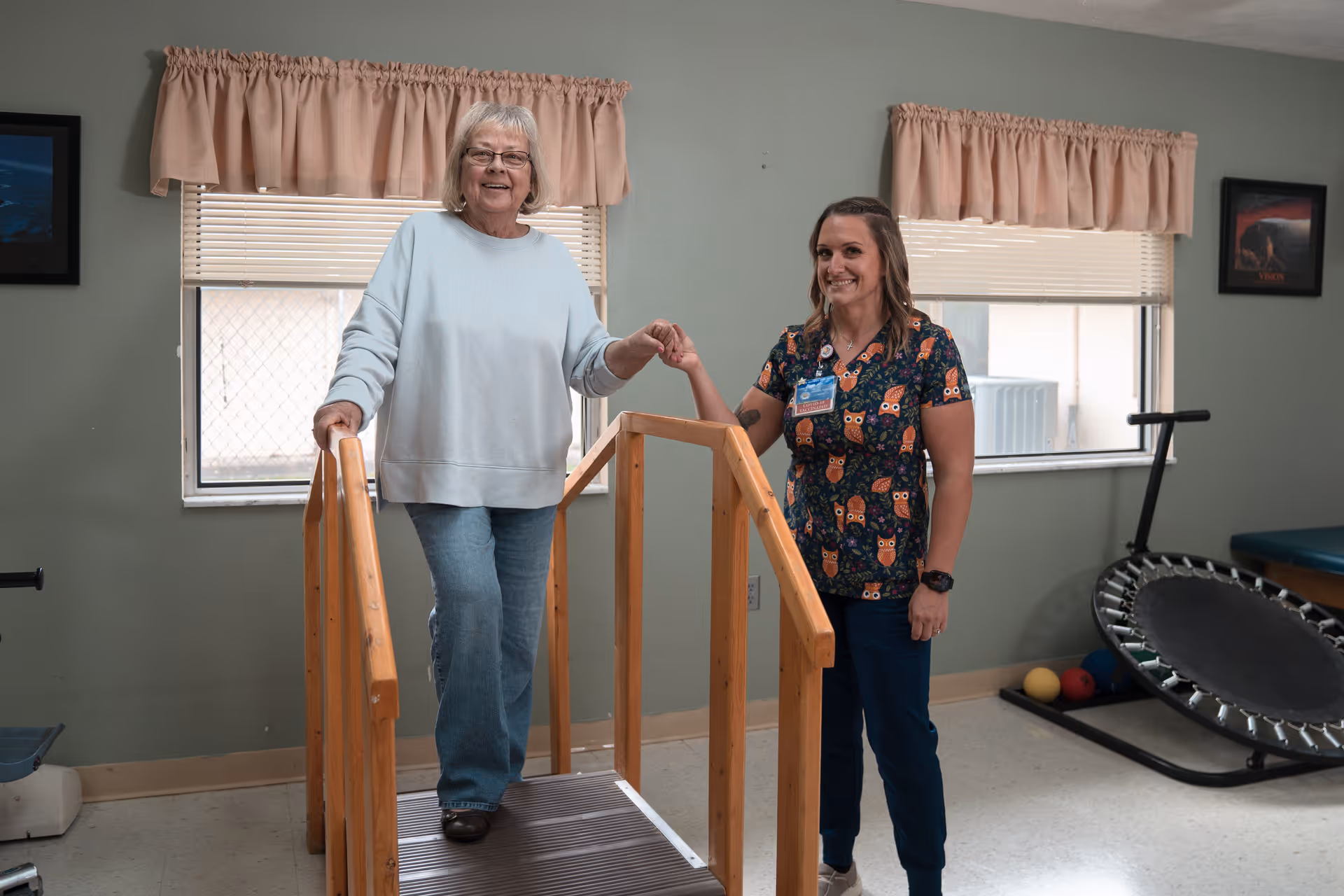 An elderly woman is walking up a small wooden ramp inside a room, assisted by a smiling female caregiver wearing a colorful owl-patterned scrub top. The room has two windows with beige curtains, a small trampoline, and some exercise balls in the background.
