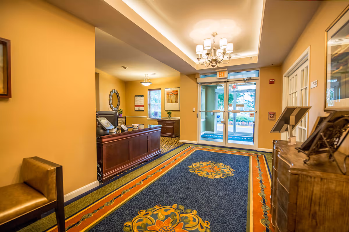 Interior view of a senior living facility entrance area with a decorative blue and orange carpet, a wooden reception desk, a chandelier, framed artwork on the walls, and glass double doors leading outside.