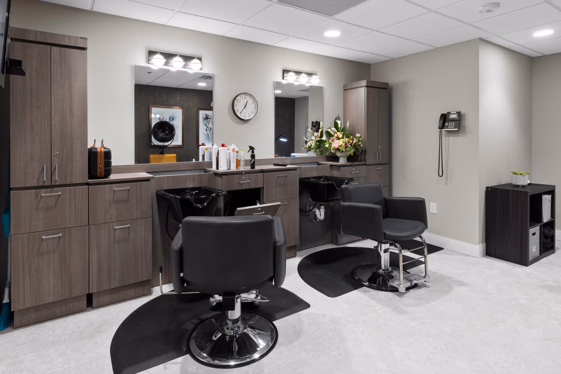 Interior view of a senior living facility's hair salon area featuring two black salon chairs in front of a counter with two sinks, large mirrors, and various hair care products. The space has modern cabinetry, a wall-mounted telephone, a clock, and a small shelving unit with decorative plants.