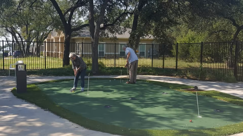Two older adults putting on a small outdoor putting green next to a fenced lawn and trees with a building in the background.