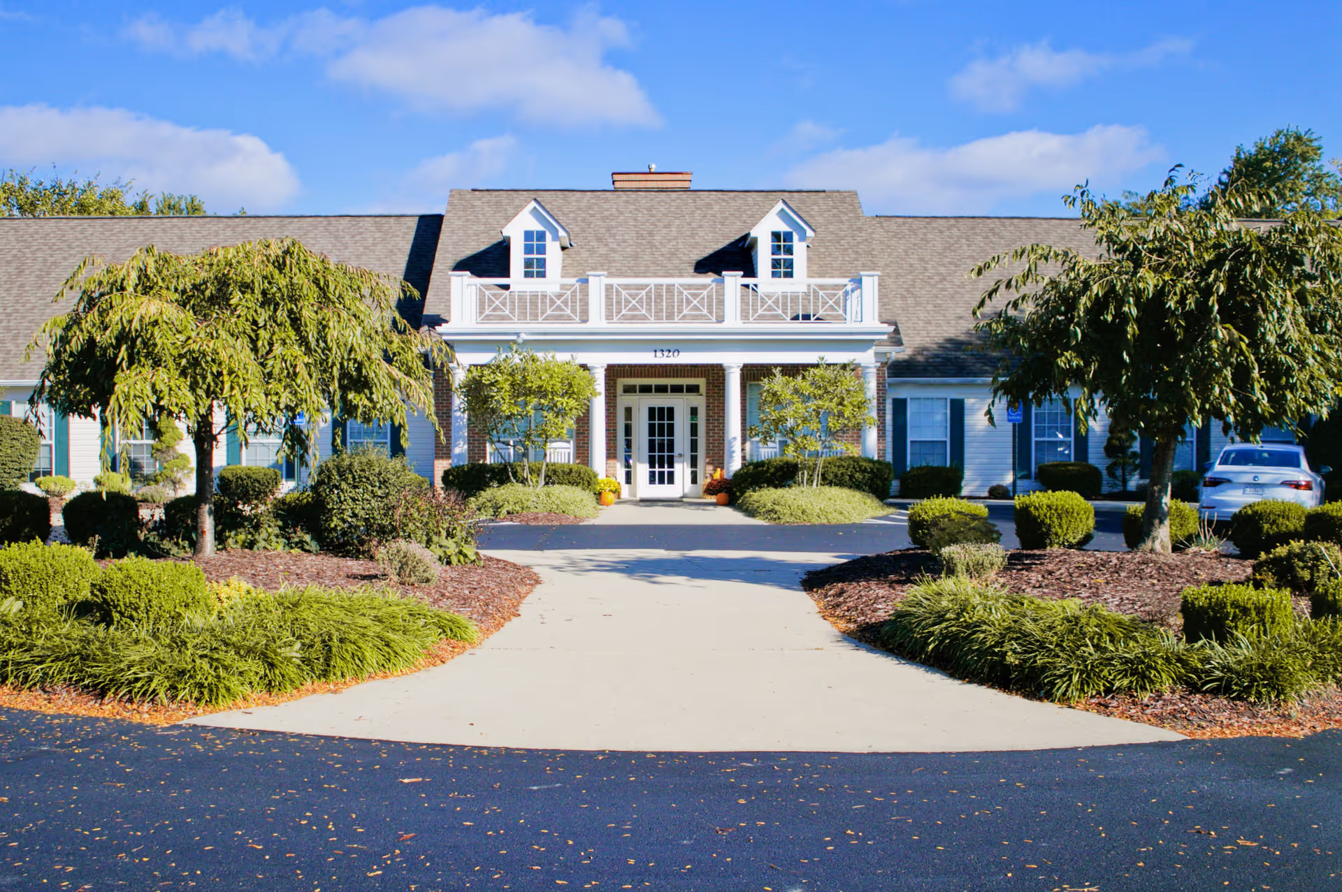 Front entrance of a single-story senior living building with a driveway, landscaped shrubs and trees, and a blue sky overhead.