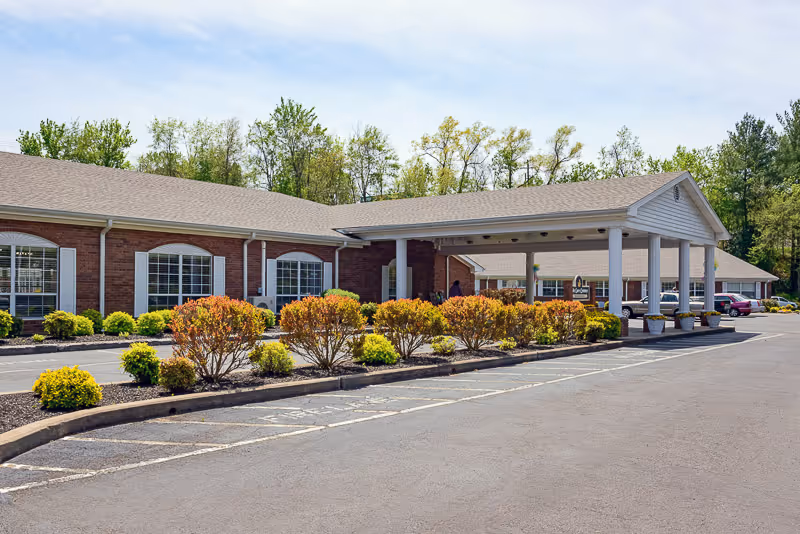 Exterior view of a single-story brick building with white trim and a covered entrance supported by white columns. There are neatly trimmed bushes and small trees along the front, a paved parking area, and a backdrop of tall green trees under a partly cloudy sky.