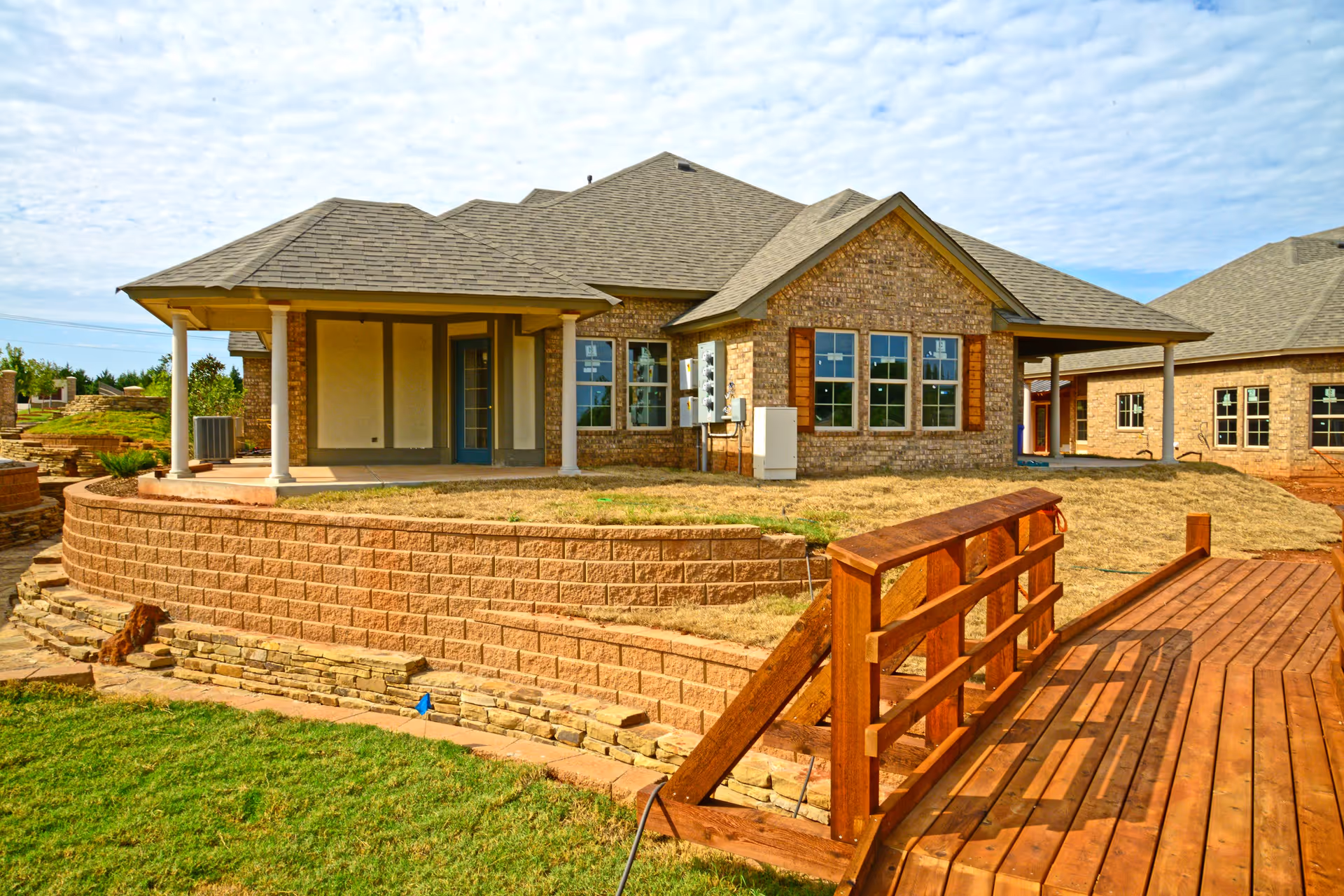Exterior view of a brick building with a covered porch supported by white columns, multiple windows with wooden shutters, and a wooden bridge leading to the entrance. The building is surrounded by a retaining wall made of stone blocks and a grassy area.