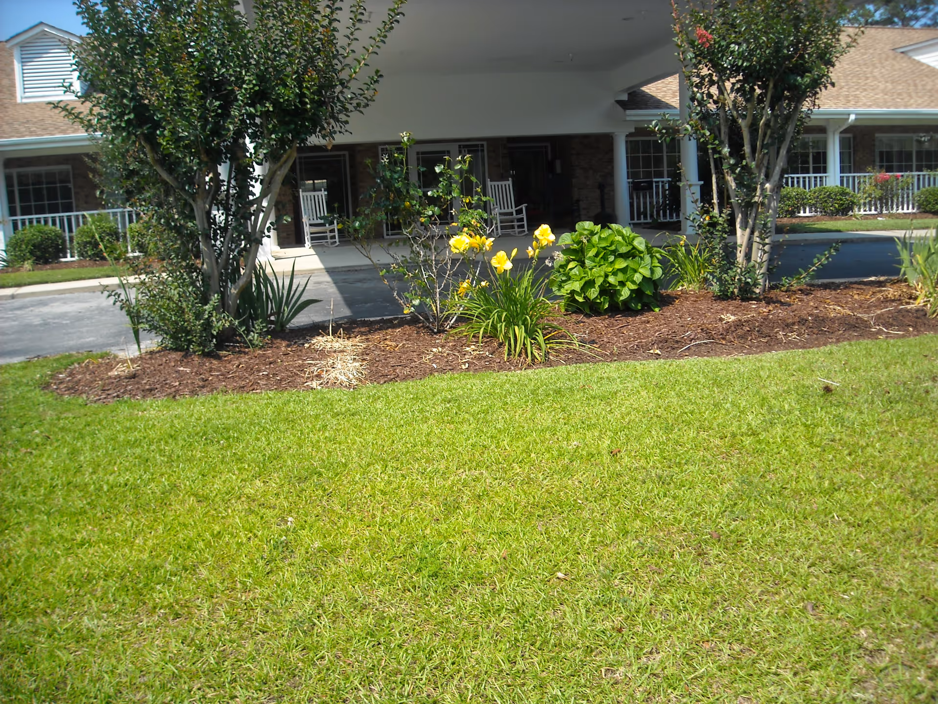 Front exterior view of Carteret House showing a well-maintained garden with green grass, yellow flowers, shrubs, and small trees. The building has a covered entrance with white rocking chairs on the porch and brick walls with white trim.
