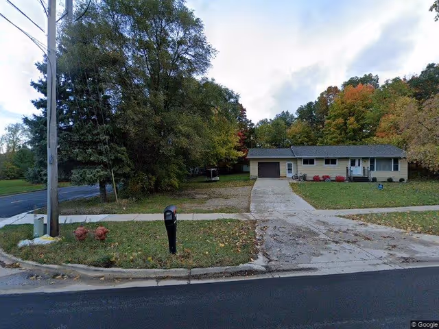 A single-story house with a driveway and garage, surrounded by trees with autumn foliage. There is a mailbox and two fire hydrants near the curb, and a sidewalk runs in front of the house.