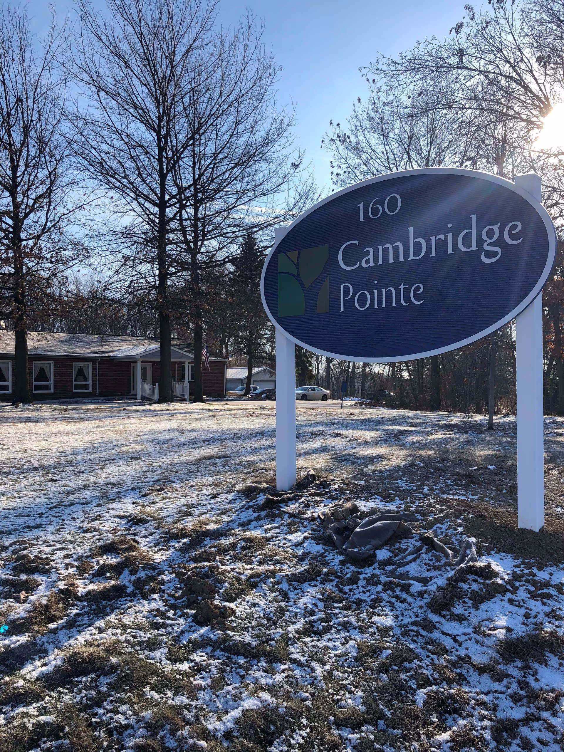 Outdoor scene showing a large oval sign with the text '160 Cambridge Pointe' in front of a building. The ground is partially covered with snow and there are leafless trees around. The sun is shining from the right side of the image.