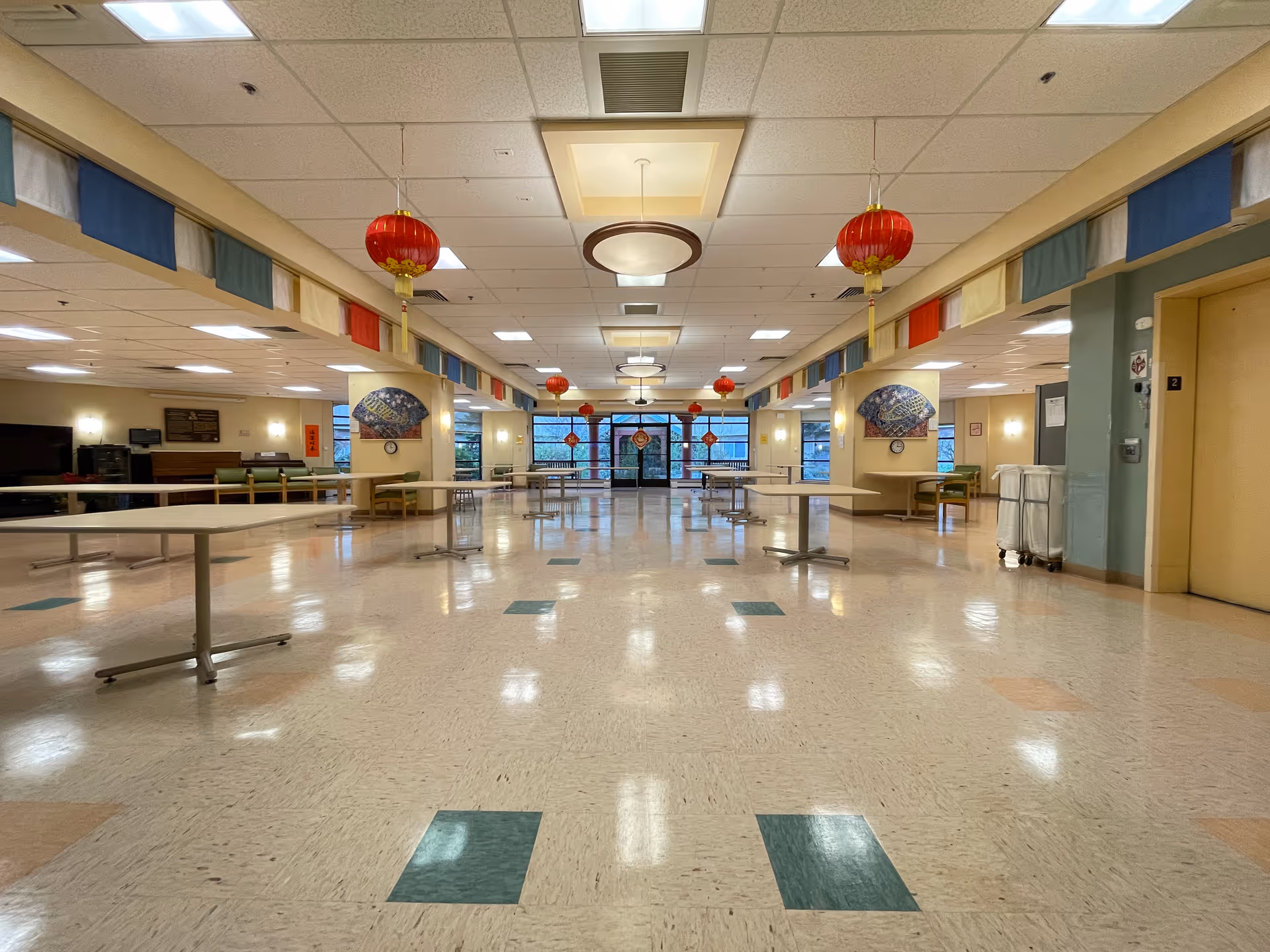 A large, well-lit common area with a polished floor featuring green and beige tiles. Several round tables with metal legs are spaced throughout the room. Red hanging lanterns and colorful fabric banners decorate the ceiling. The walls have artwork and windows, and there is an elevator on the right side. The room appears clean and spacious with a welcoming atmosphere.