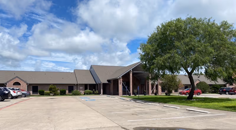 Single-story brick nursing and rehabilitation building with a covered main entrance, adjacent parking lot, and a large tree under a partly cloudy sky.