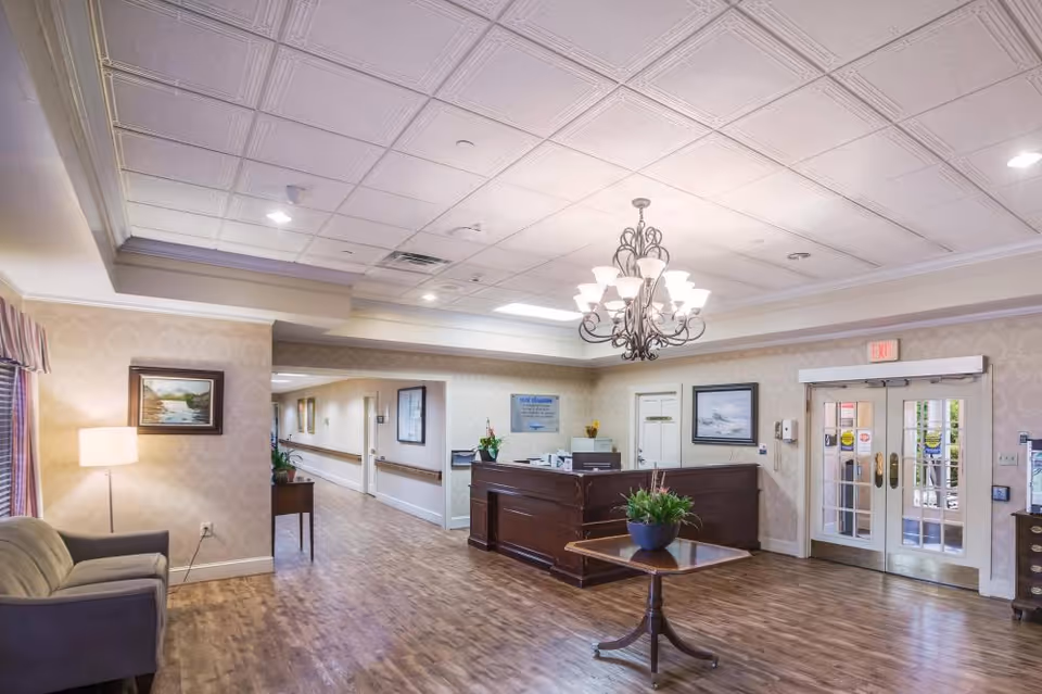 Reception area of a senior living facility with a wooden reception desk, a chandelier hanging from the ceiling, a small table with a plant, a couch with a floor lamp, framed paintings on the walls, and double glass doors leading outside.