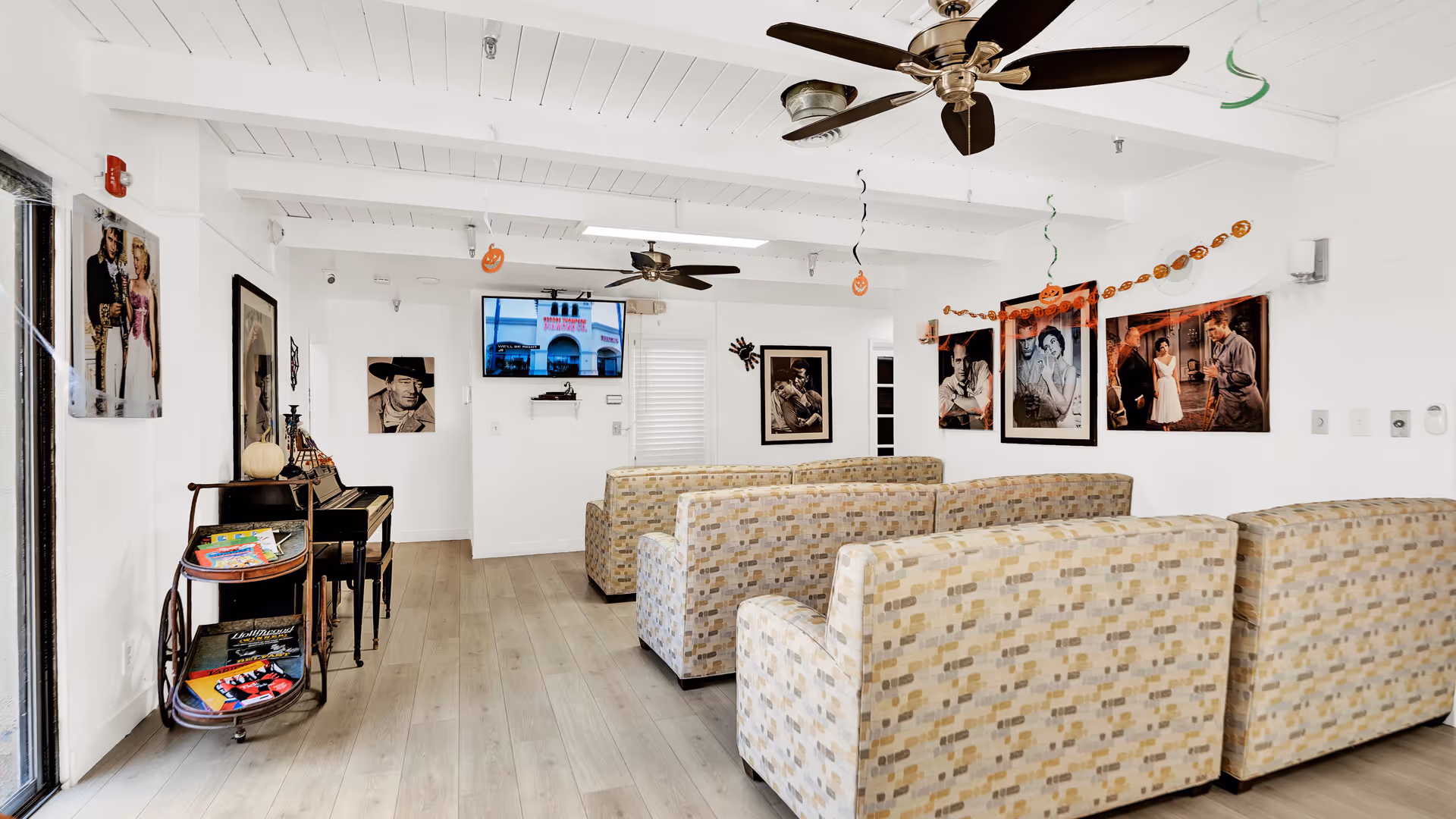 Interior of a senior living facility lounge with patterned armchairs arranged in rows facing a wall-mounted TV. The room has white walls decorated with black and white movie posters and Halloween-themed hanging decorations. There is a piano and a small cart with magazines on the left side. The ceiling has exposed beams and two ceiling fans.