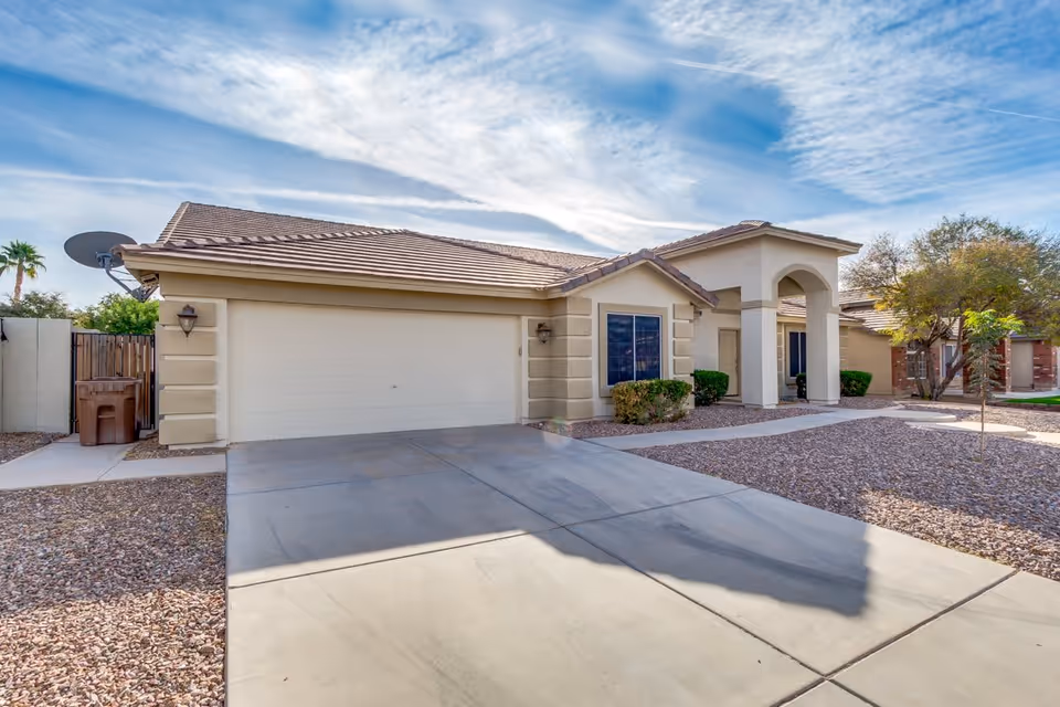 Exterior view of a single-story residential building with a two-car garage, beige stucco walls, and a tiled roof under a partly cloudy sky. The driveway and front yard are covered with gravel and there are small bushes near the entrance.