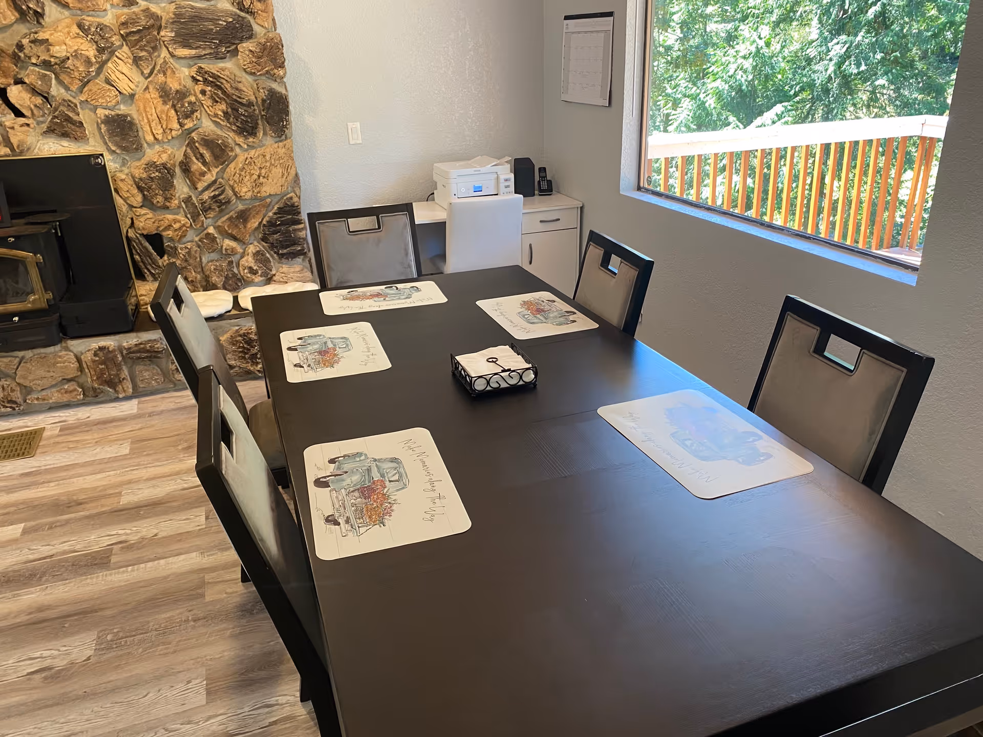 A dining area with a dark wooden table set with six chairs, each with a placemat featuring a vintage truck design. There is a stone fireplace on the left side and a large window on the right showing green trees outside. In the corner, there is a small white desk with a printer and a phone on it.