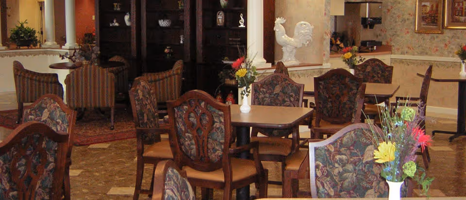 Interior view of a dining area with multiple wooden tables and chairs featuring floral upholstery. Each table has a small vase with colorful flowers. In the background, there is a dark wooden cabinet with decorative items and a white rooster sculpture on a column. The room has warm lighting and a cozy atmosphere.