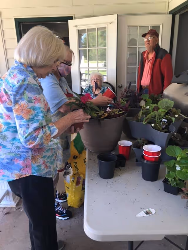 A group of elderly people and a caregiver are gathered around a table on a covered porch, engaging in a gardening activity with various potted plants and gardening supplies on the table.