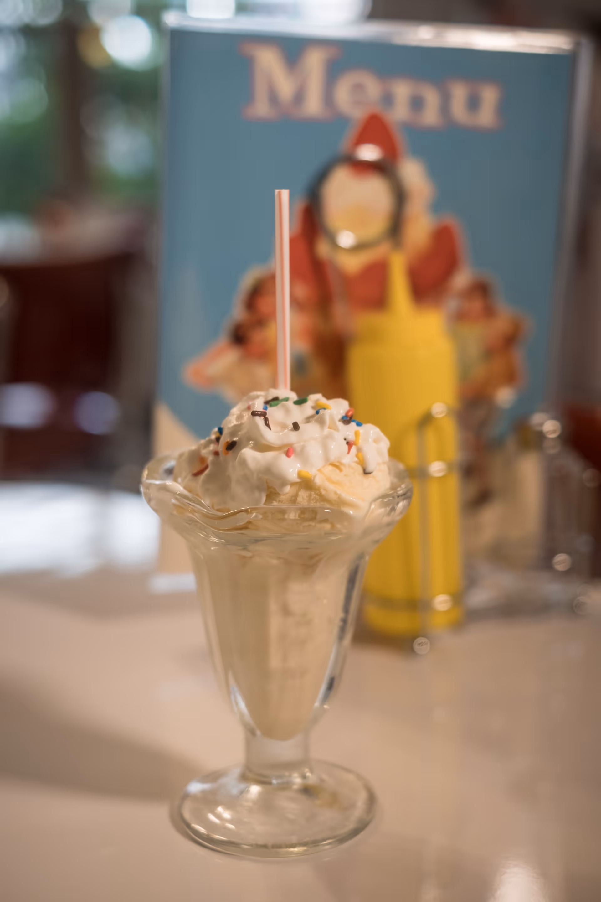 A glass sundae dish filled with vanilla ice cream topped with whipped cream and colorful sprinkles, with a straw inserted. In the background, there is a blue menu card and a yellow mustard bottle on a table in a dining setting.