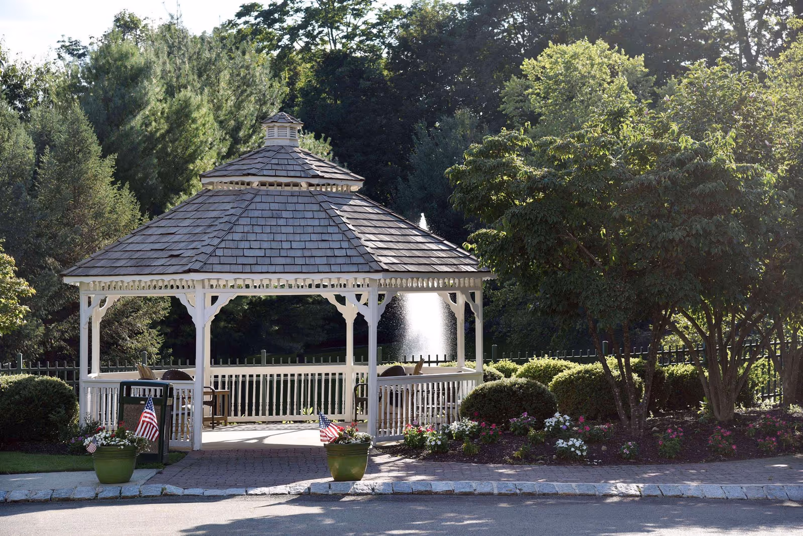 A white wooden gazebo with a shingled roof situated in a landscaped garden area with green bushes, flowering plants, and trees. There are two green flower pots with small American flags on either side of the gazebo entrance. In the background, a water fountain is visible surrounded by a black metal fence and dense trees.