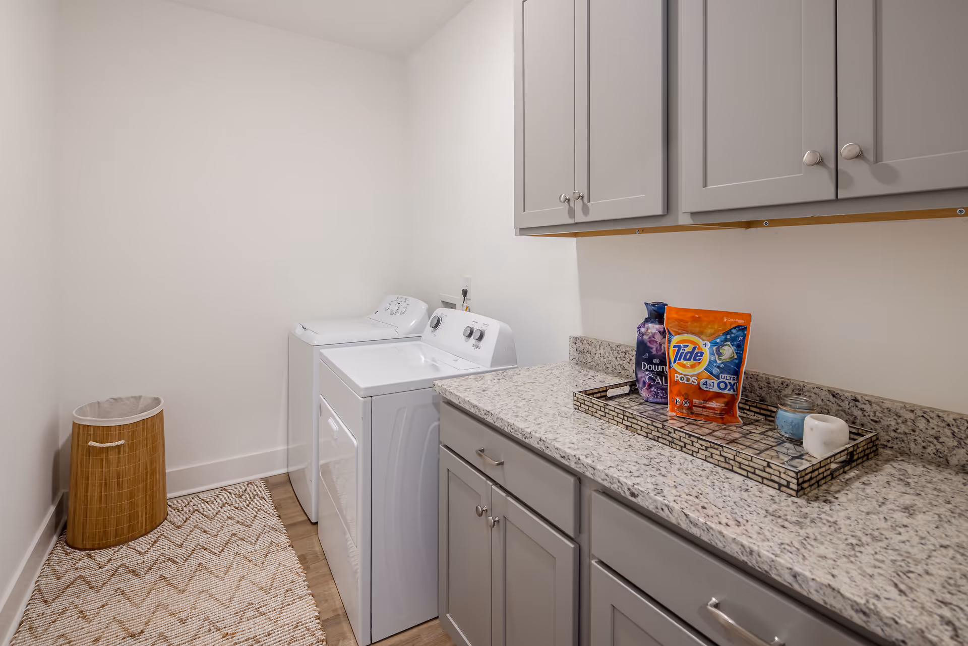 Laundry room with a white washing machine and dryer side by side, gray cabinets above and below a granite countertop, a woven laundry basket in the corner, and a tray on the counter holding a bottle of Downy fabric softener, a bag of Tide Pods, and a small candle.