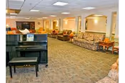 Spacious common area with patterned carpet, multiple sofas and armchairs arranged along the walls, a black piano with a bench in the foreground, and warm lighting from ceiling fixtures.