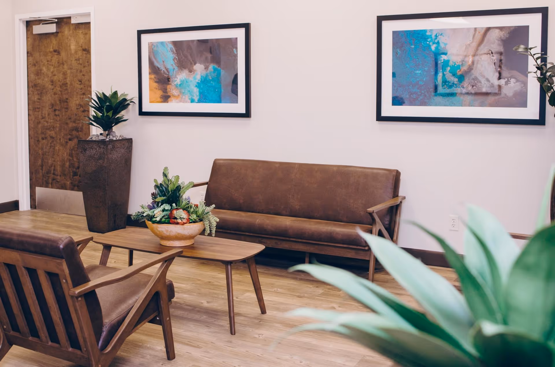A cozy seating area with a brown leather sofa and matching armchair around a wooden coffee table with a decorative plant centerpiece. The room has light-colored walls adorned with two abstract paintings in black frames. There is a wooden door and a tall planter with a green plant in the corner. Another green plant is partially visible in the foreground.